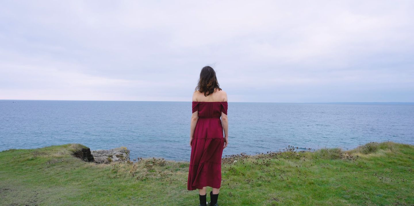 Woman in burgundy off-shoulder dress standing on grassy cliff edge overlooking calm blue ocean under overcast sky, seen from behind.