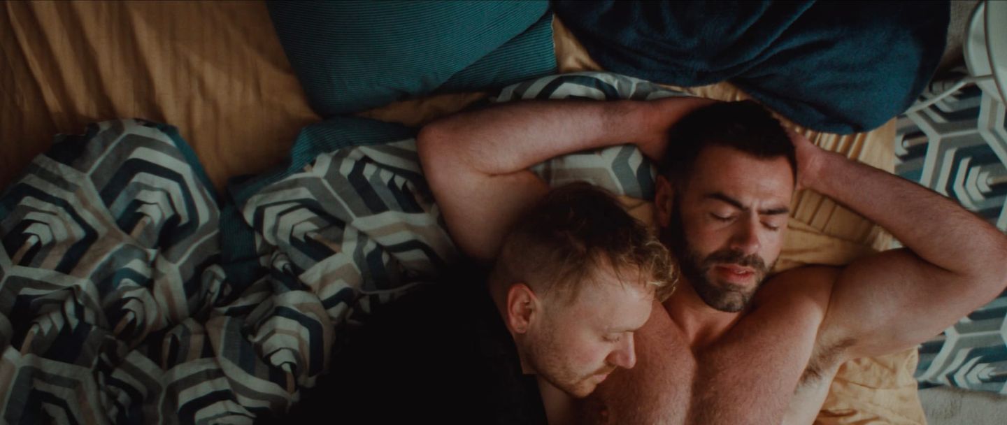 Two shirtless men sleeping closely together in bed, viewed from above, surrounded by geometric-patterned and teal bedding.