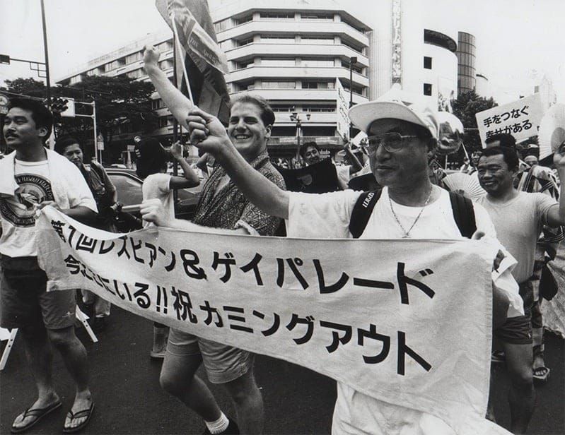 Black and white photo of marchers at a Japanese Lesbian & Gay Pride Parade holding a banner with Japanese text celebrating coming out.