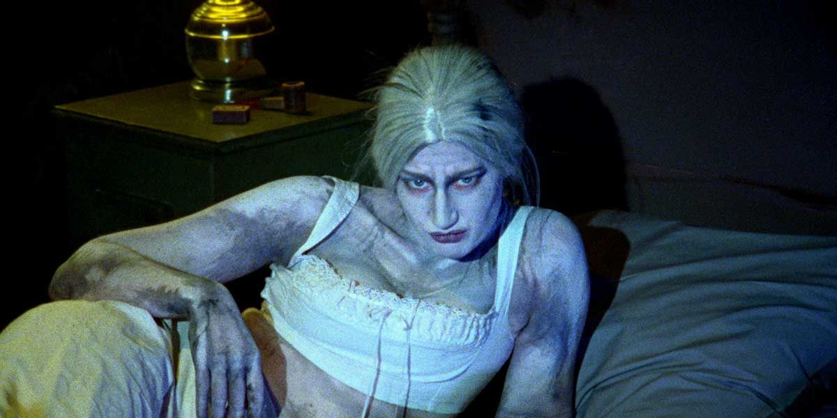 Gaunt figure with pale bluish skin and white hair in a nightgown, sitting on a bed in a dark room, from a horror film.