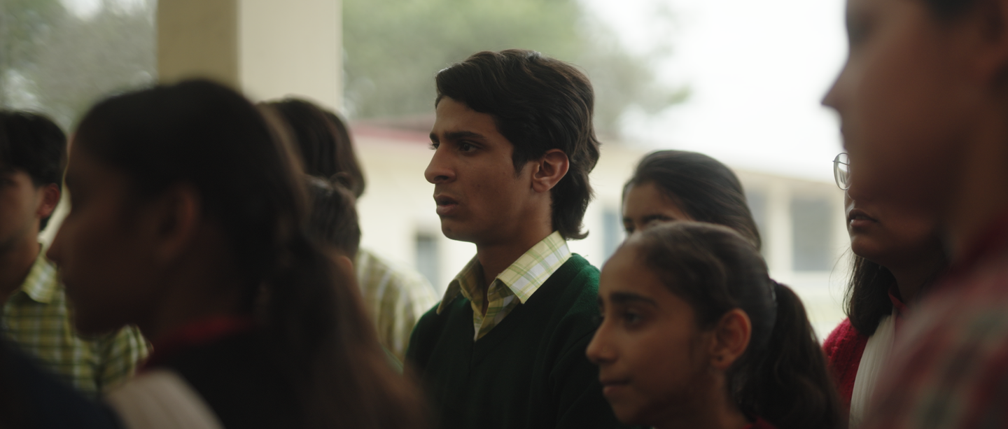 Young man in green sweater over plaid shirt stands among a crowd of students in what appears to be an Indian school setting.