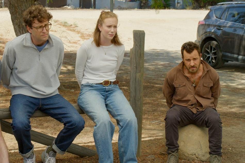 Three people sitting outdoors on a wooden fence and rock; two men and a woman in casual clothes, with a parked SUV behind them.