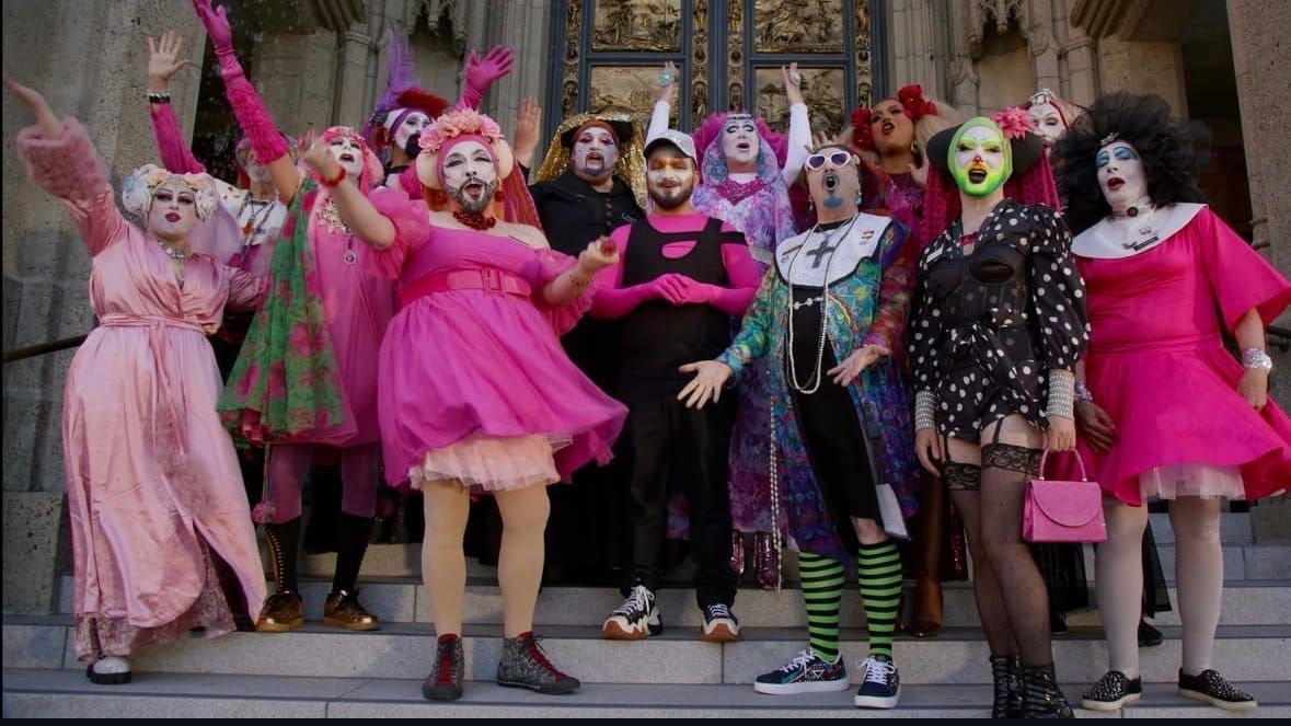 Group of drag performers in colorful costumes and theatrical makeup posing on stone steps in front of ornate golden doors.