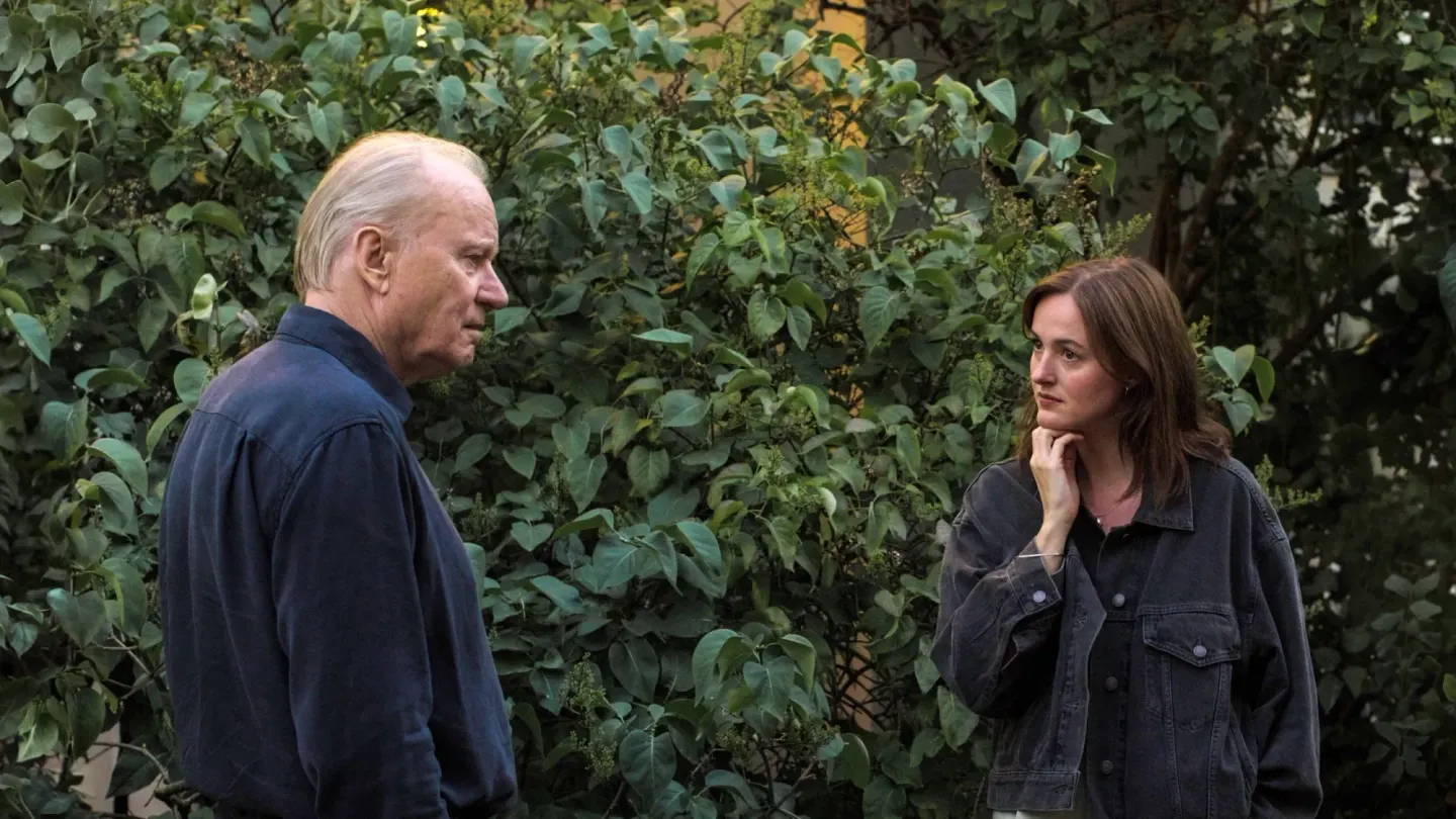 An older man in a navy shirt faces a younger woman in a dark denim jacket, talking outdoors near a lush green hedge.