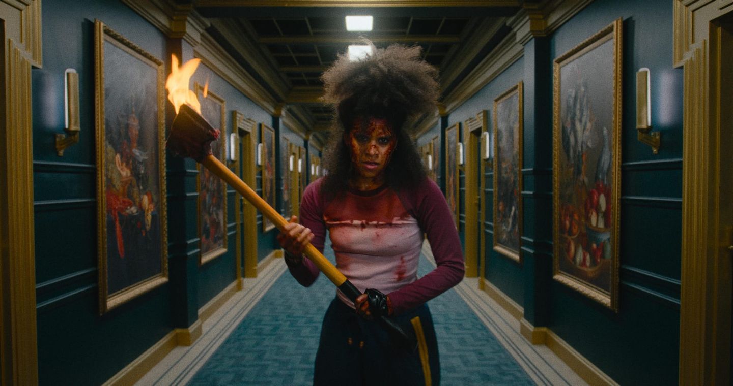 Blood-covered woman holding a flaming torch in an ornate hallway lined with gold-framed paintings, looking determined.