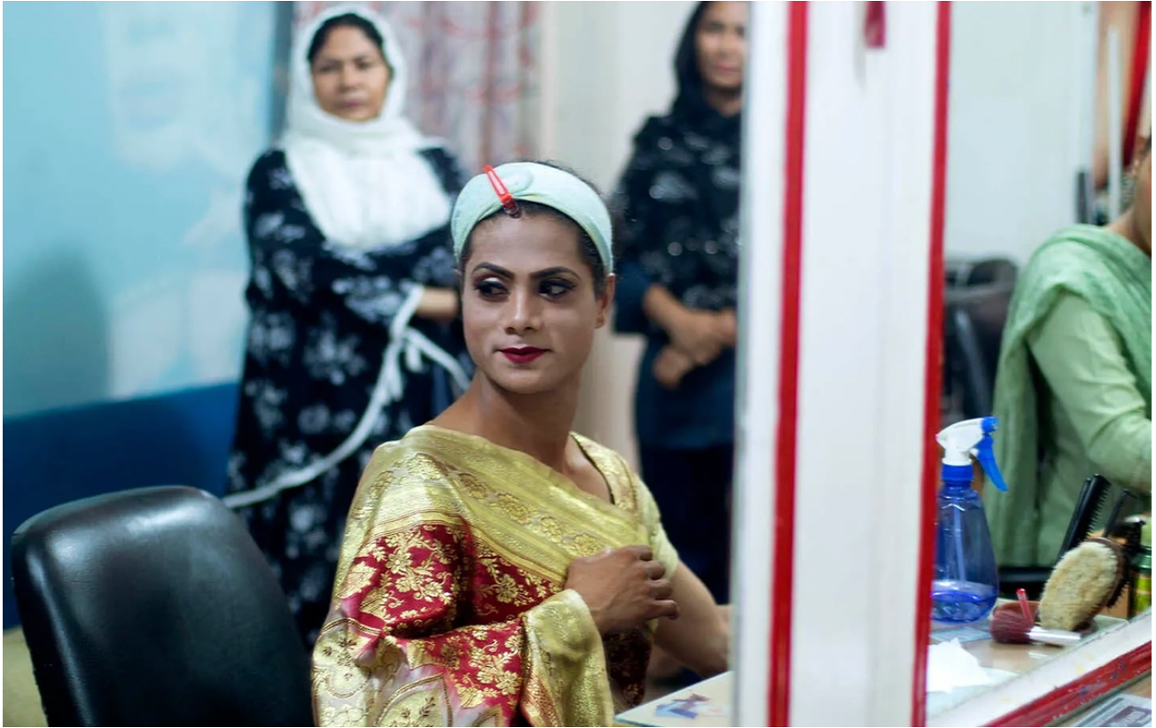 Person in gold and red saree getting makeup done at a beauty salon, with headband holding hair back, another woman standing behind.