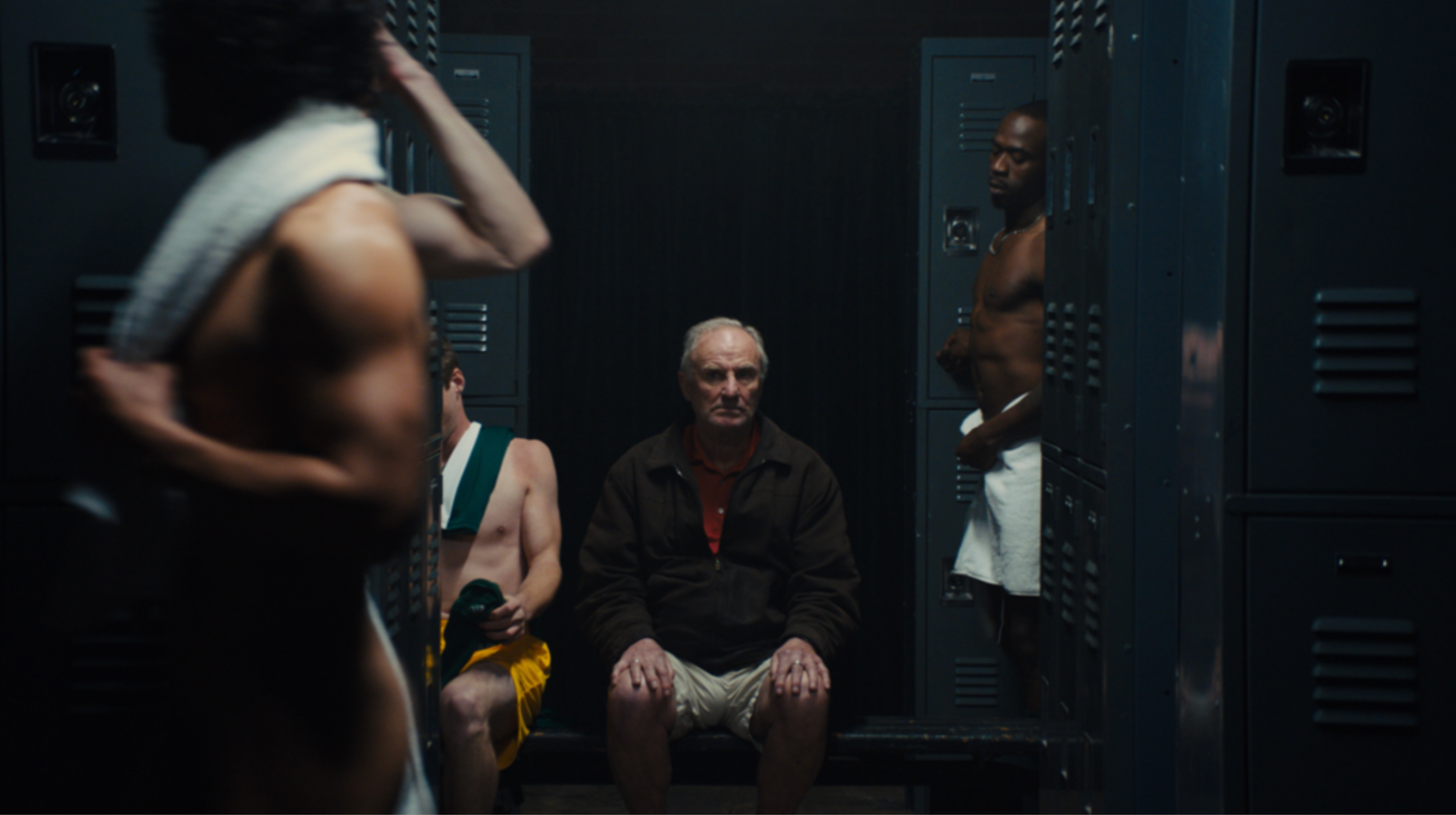 Older man sitting in a dimly lit locker room surrounded by younger athletes changing, with blue metal lockers behind them.