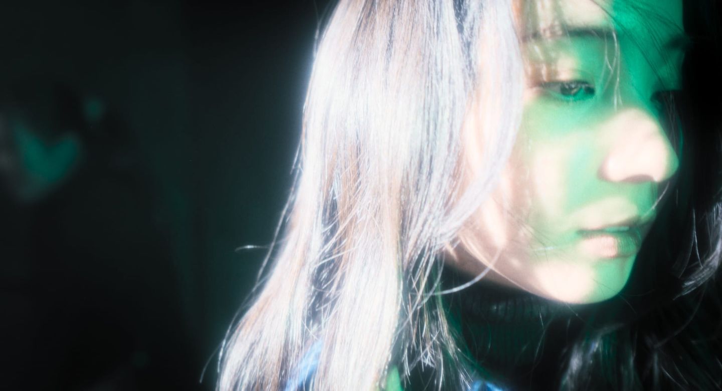 Close-up of a young woman's face partially obscured by blonde-streaked hair, illuminated by green light against a dark background.