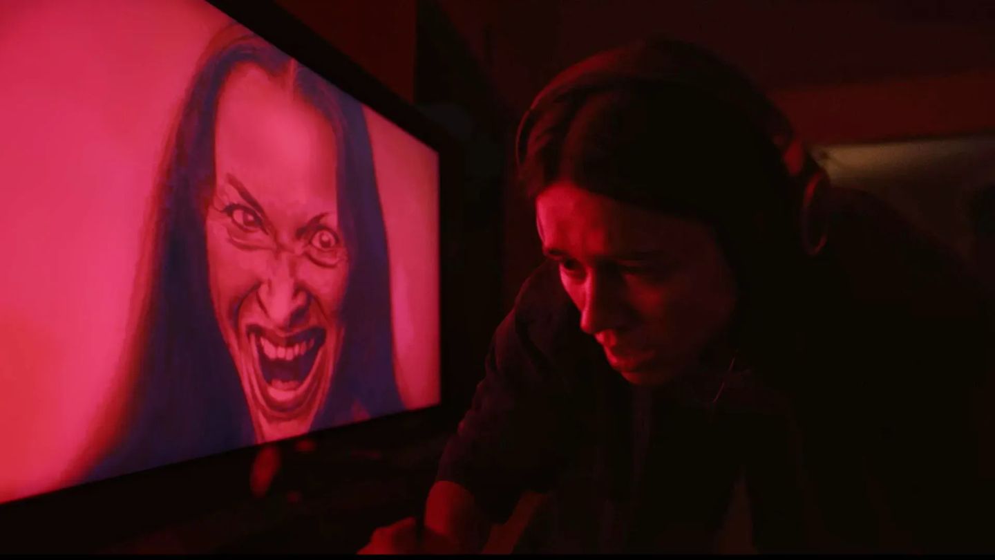 Person in red-lit room next to a screen displaying a disturbing drawing of a woman with wide eyes and a menacing grin.