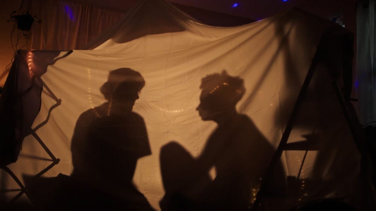 Two silhouettes of people facing each other cast on a backlit white bedsheet, with warm lighting and string lights visible.