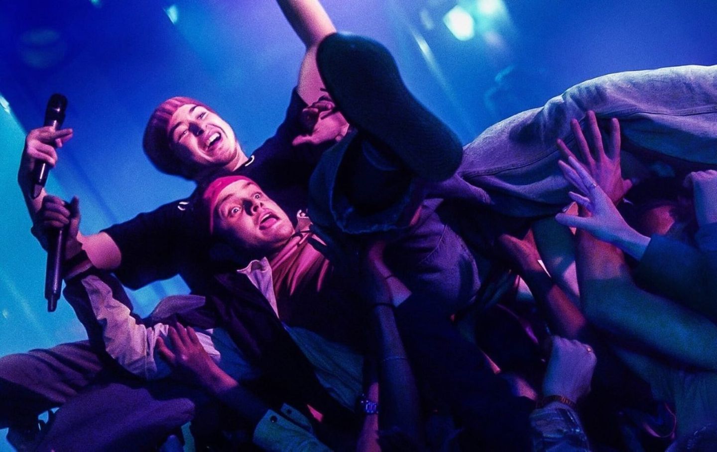 Two young men crowd-surfing at a concert under blue and purple lights, one holding a microphone, supported by raised hands.