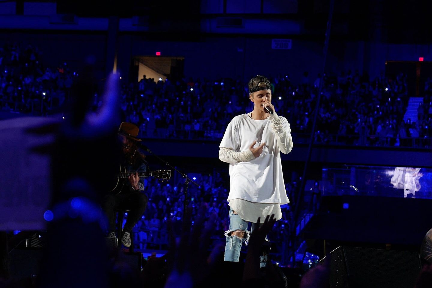 Singer in white long-sleeve shirt and ripped jeans performs on stage with microphone, guitarist nearby, packed arena behind.