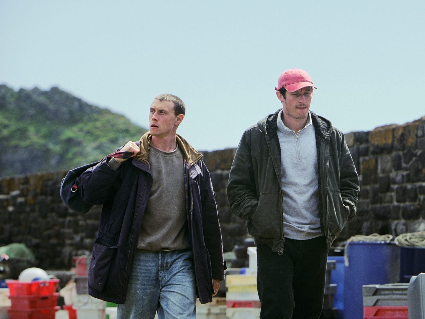 Two young men walking through a harbor area with stone walls, fishing crates, and green hills in the background.