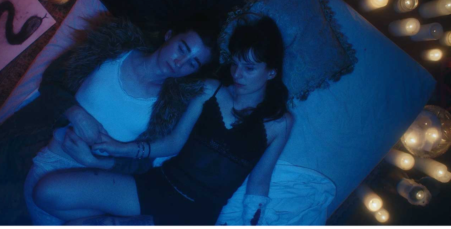 Two people lying close together on a bed bathed in blue light, surrounded by glowing candles, in an intimate nighttime scene.