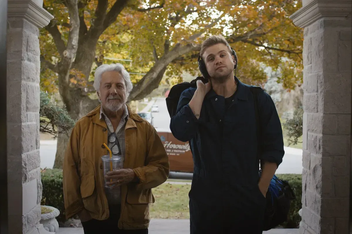 Two men on a stone porch: an older man with white hair in a tan jacket holding an iced drink, and a younger blond man with headphones around his neck carrying a backpack.