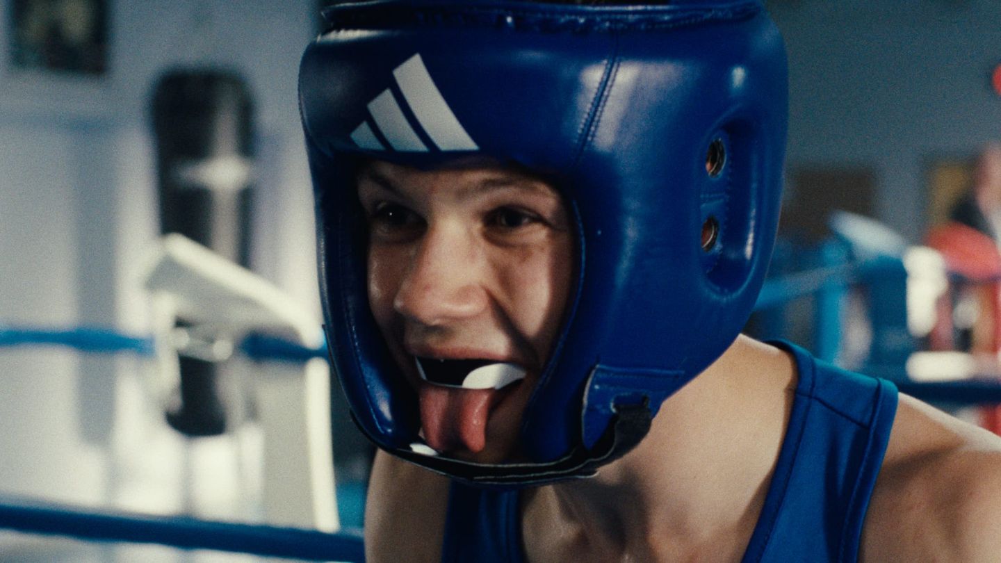 Boxer wearing blue Adidas headgear and mouthguard sticks out their tongue in a boxing ring, wearing a blue tank top.