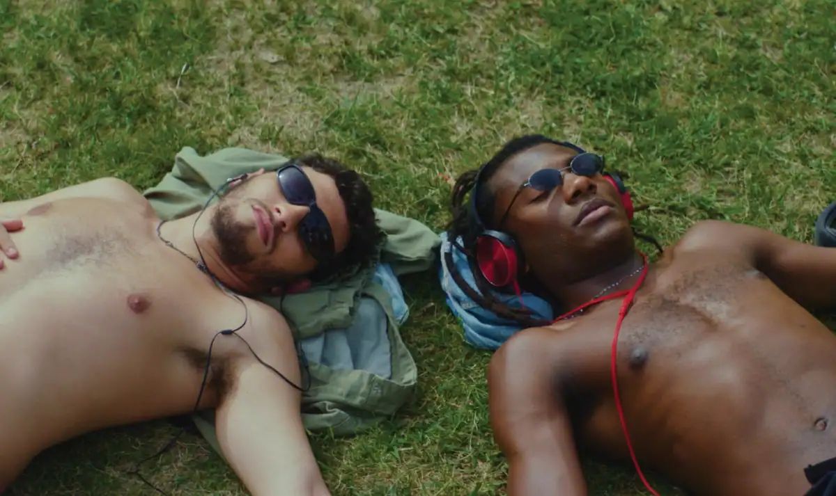 Two shirtless men lying on grass wearing sunglasses, one with headphones around his neck, sunbathing side by side.