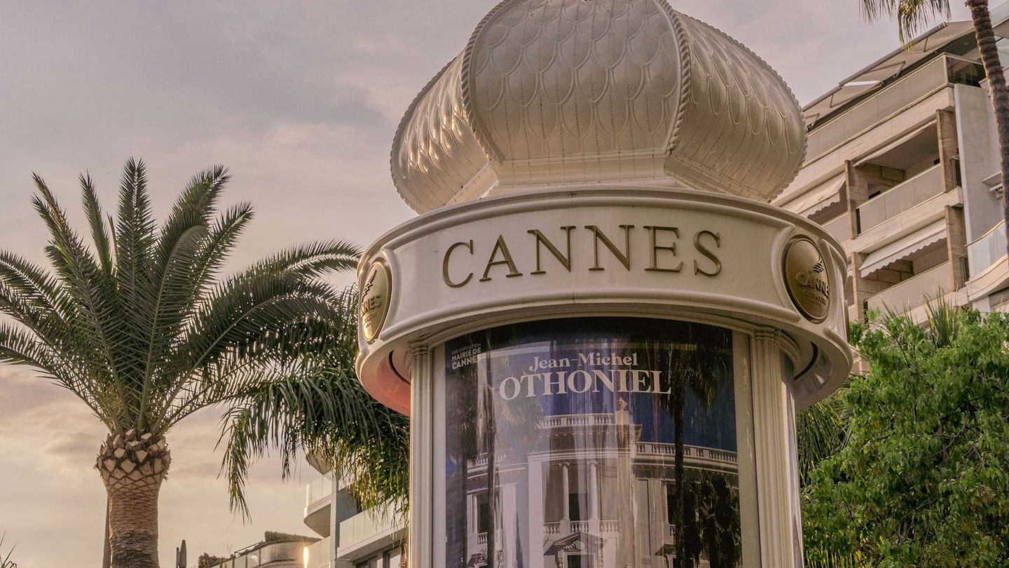 Ornate white kiosk with domed top labeled "CANNES" displaying a Jean-Michel Othoniel exhibition poster, with palm trees.