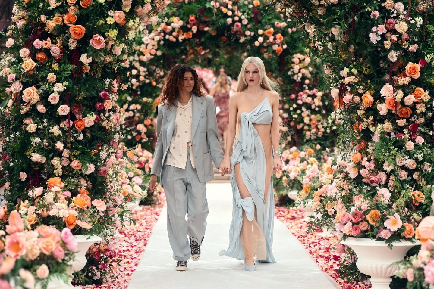 Two models walk hand-in-hand down a flower-lined runway, one in a gray suit with sneakers, the other in a blue draped gown.