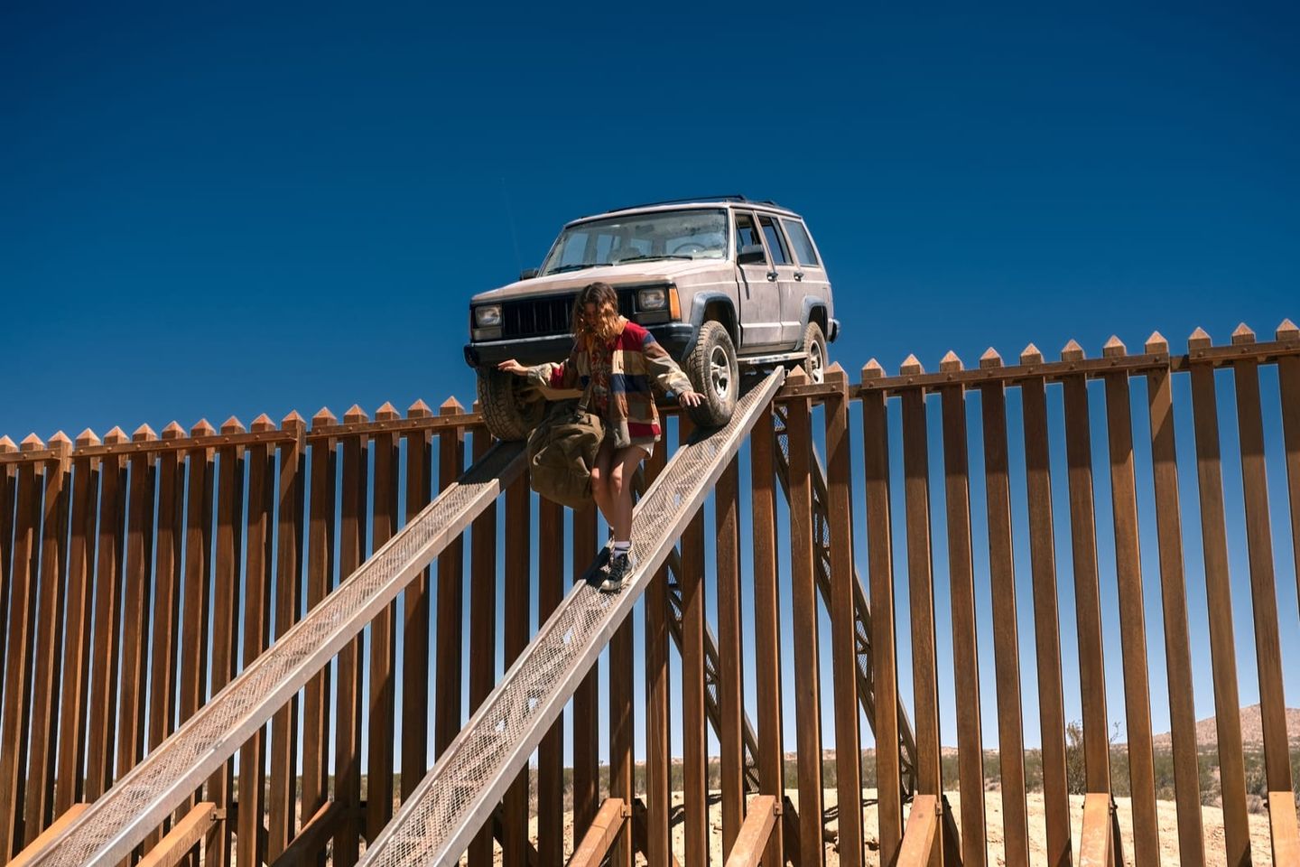 Person climbing a metal ramp over a tall brown border fence in a desert, with a Jeep Cherokee parked at the top.