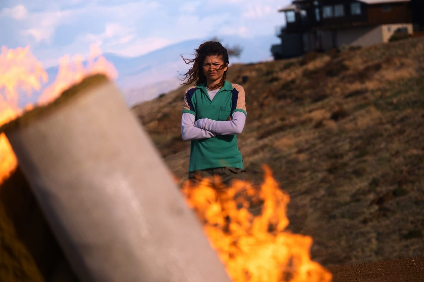 Woman with arms crossed wearing green vest and glasses stands on hillside behind burning debris, wind blowing her hair.