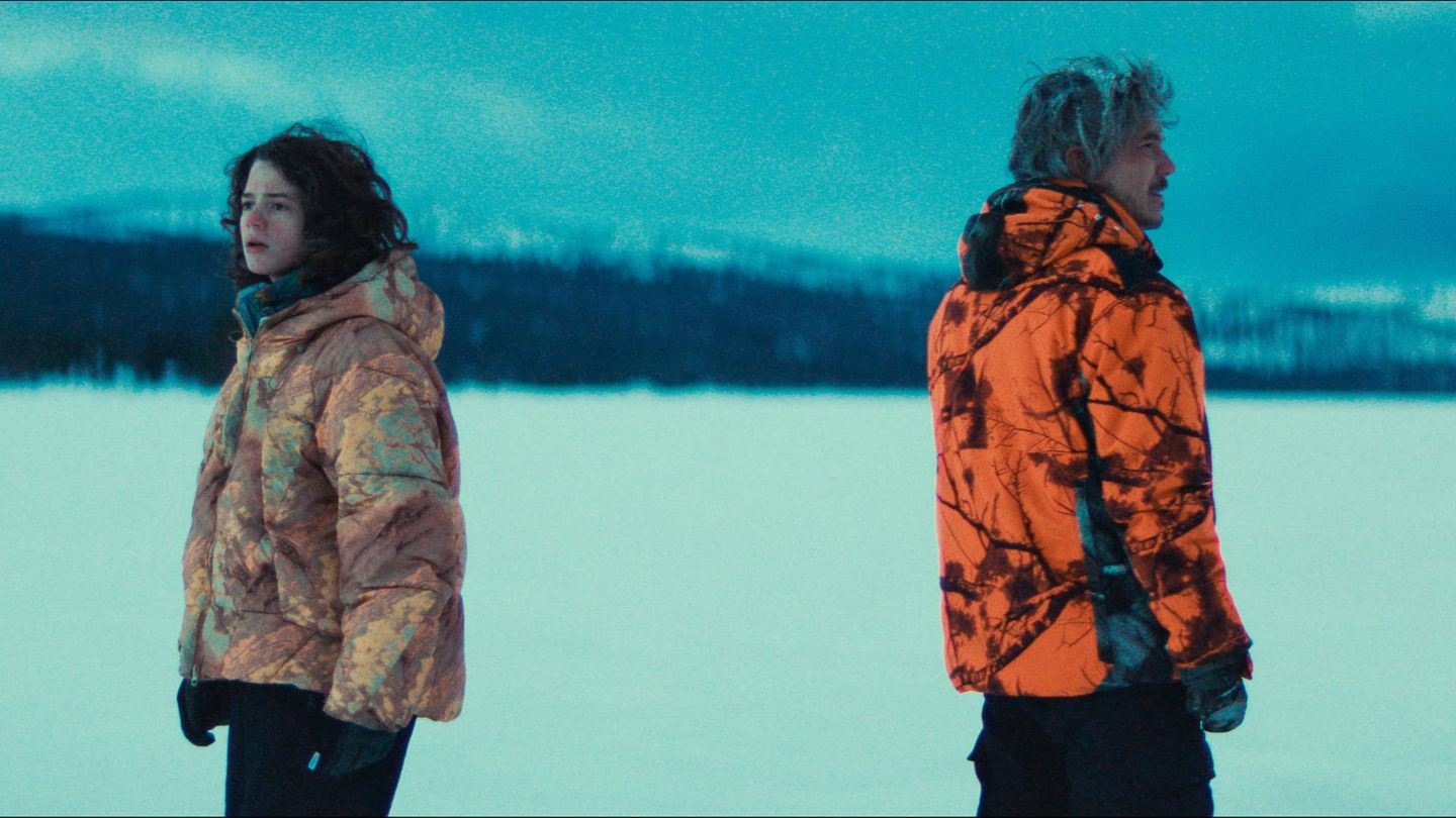 Two figures in winter jackets stand apart on a snowy island landscape.