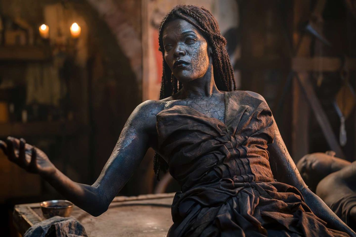 Ash-covered woman with braided hair reclines in a dim candlelit room, wearing tattered dark fabric and extending one hand.