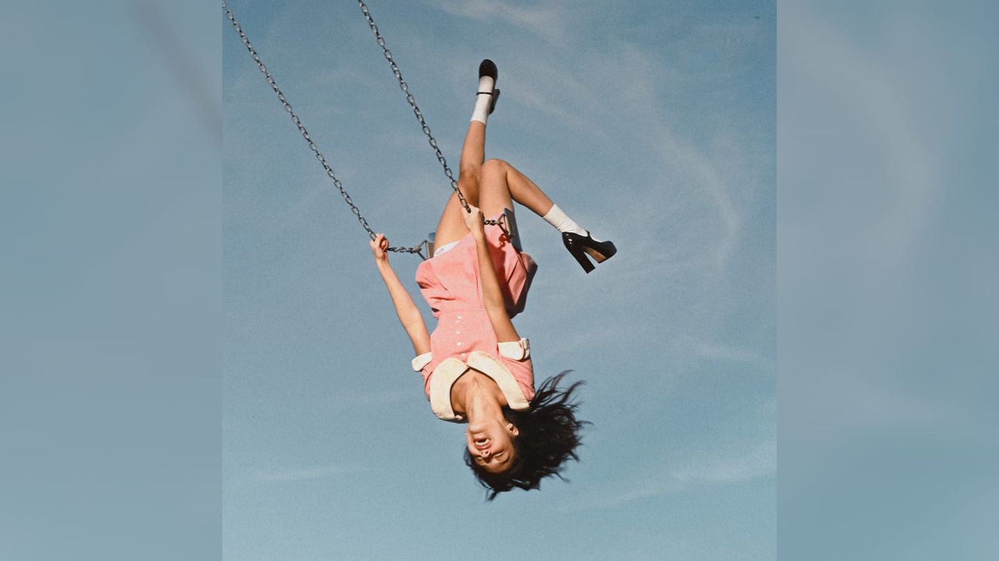 Olivia Rodrigo swings upside down in a pink outfit against a blue sky.