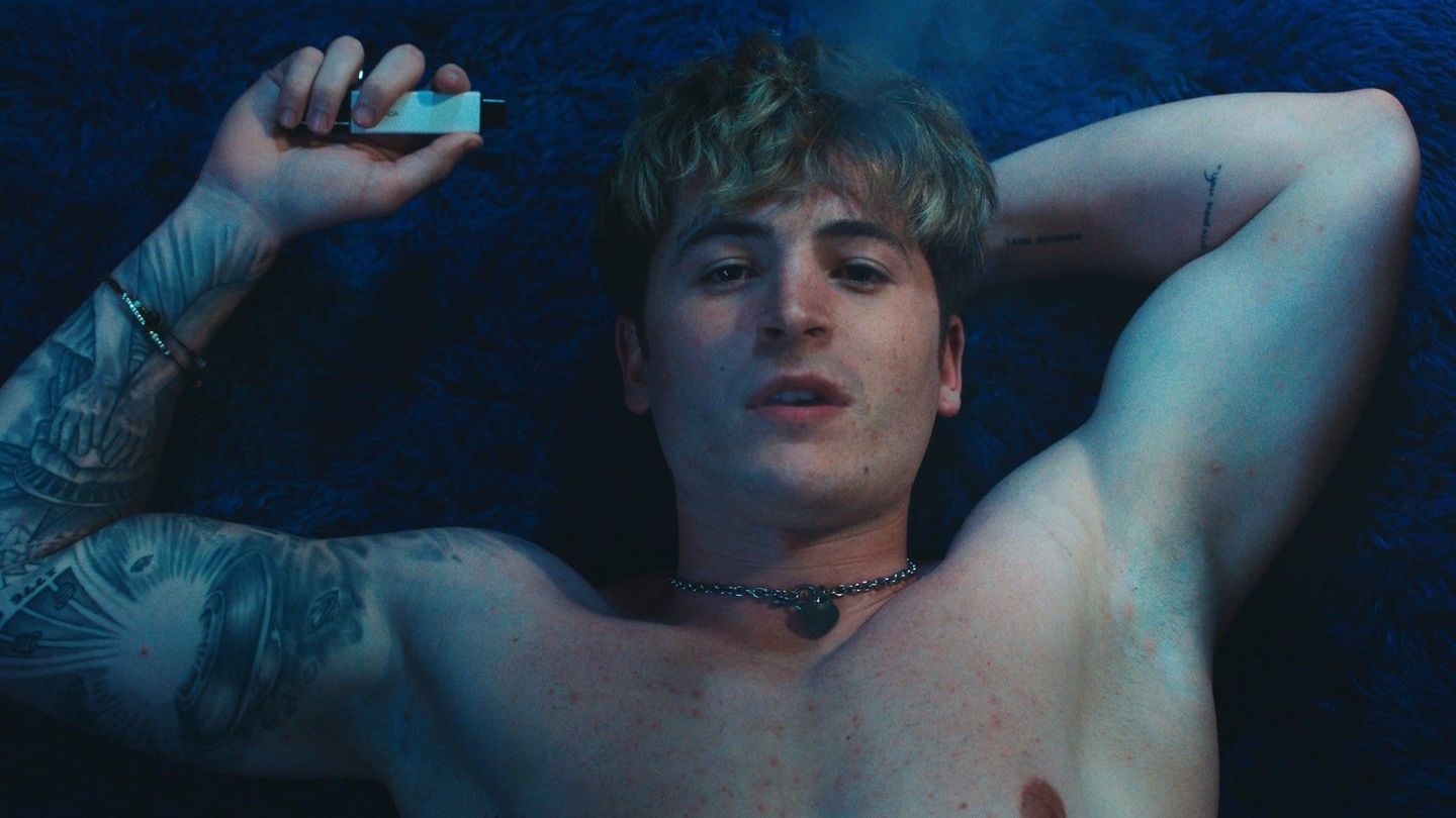 A tattooed young man lies on a dark blue bed, holding a vape.