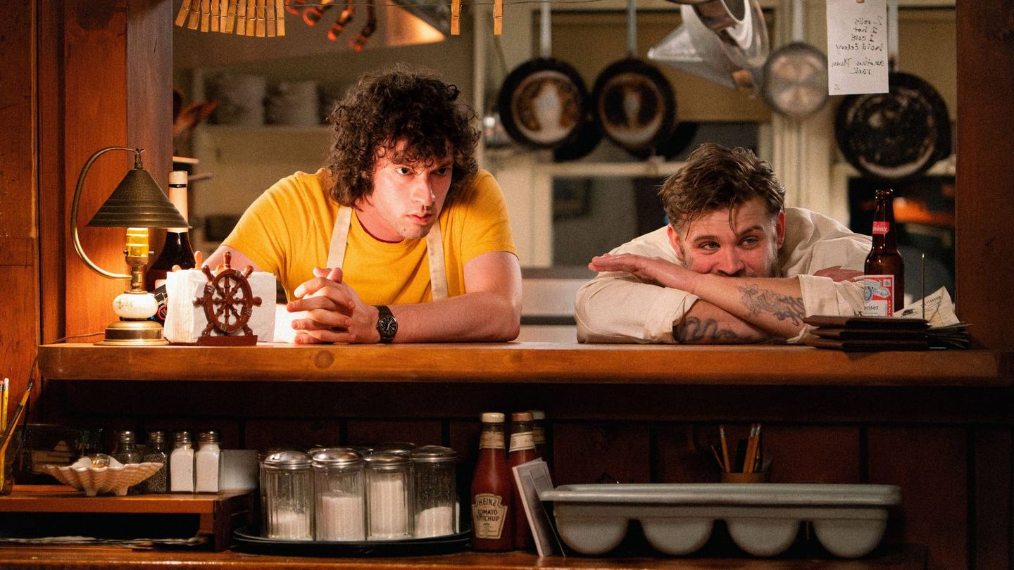 Two men lean across a diner counter in a warmly lit restaurant.