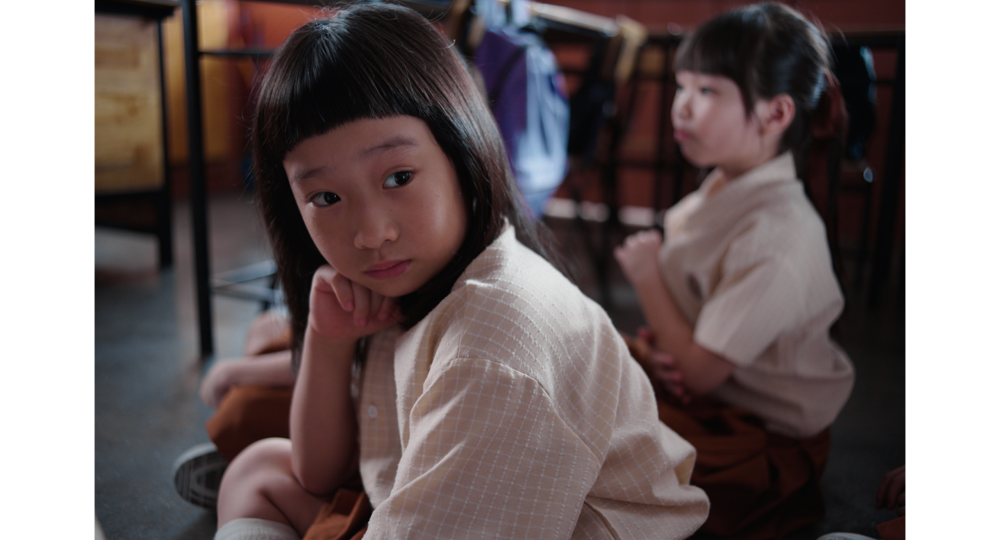 Two young girls in school uniforms sit on a classroom floor, one in the foreground and one behind her.