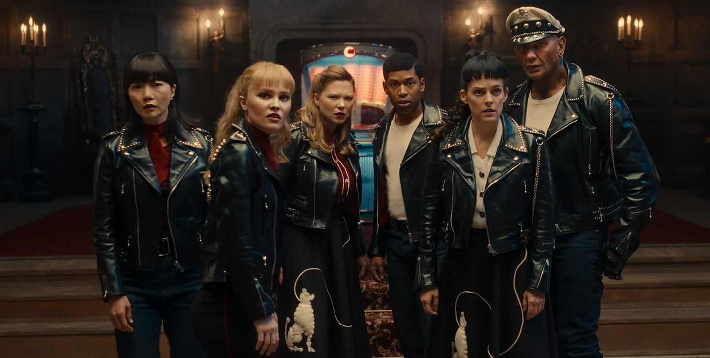 Six people in studded black leather jackets stand tensely in a dim candlelit hall, with a glowing jukebox behind them.