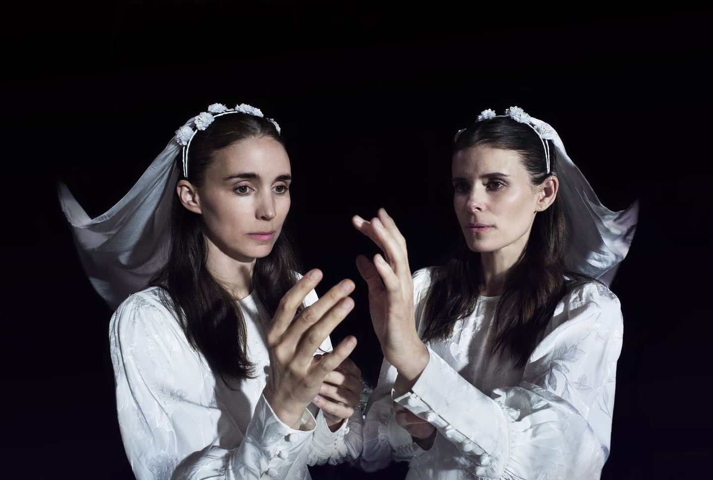 Two women in white dresses and floral veils raise their hands toward each other against a dark background.
