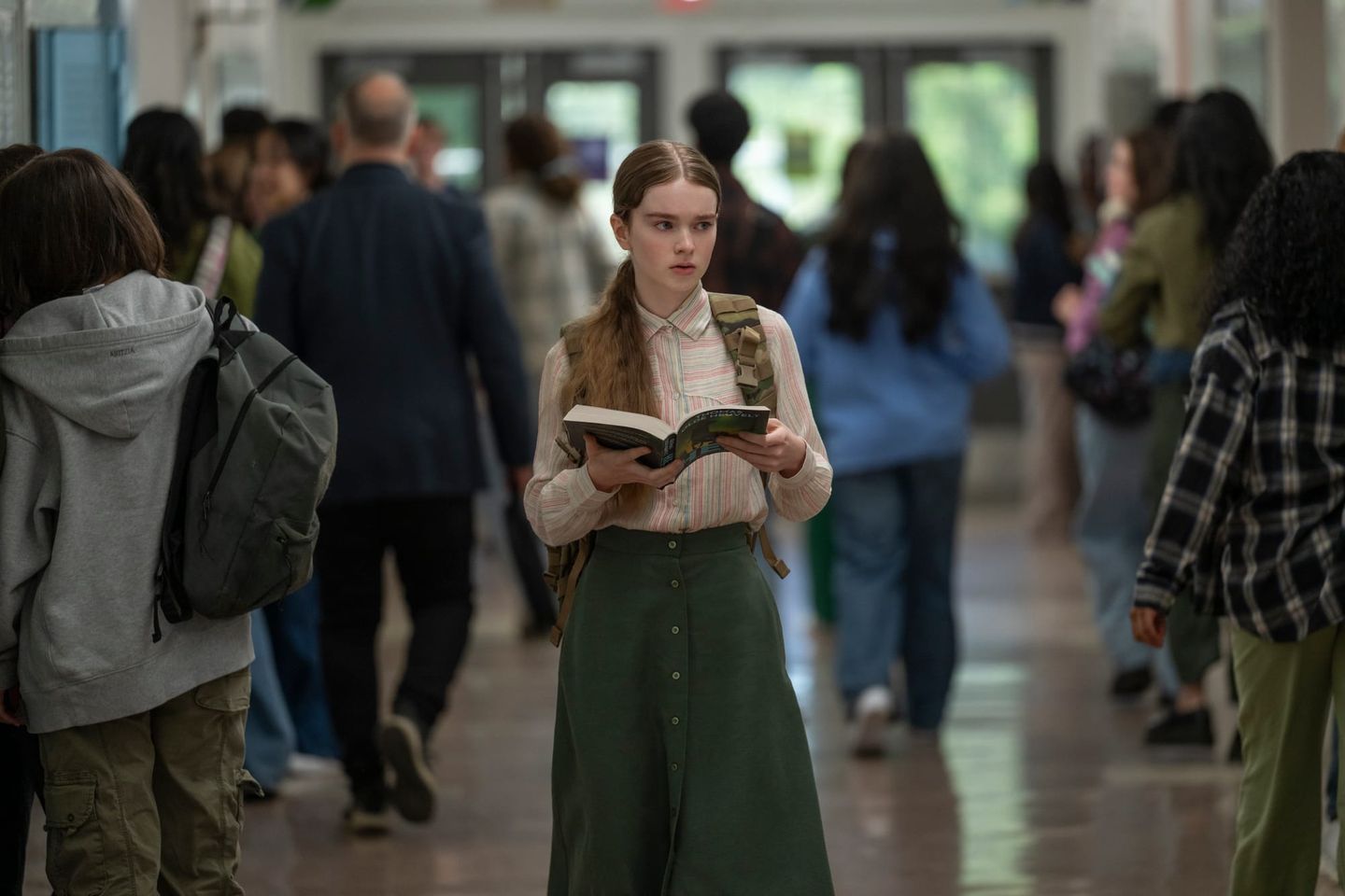 Teen girl with a backpack reads a book while standing in a crowded school hallway with students walking around her.