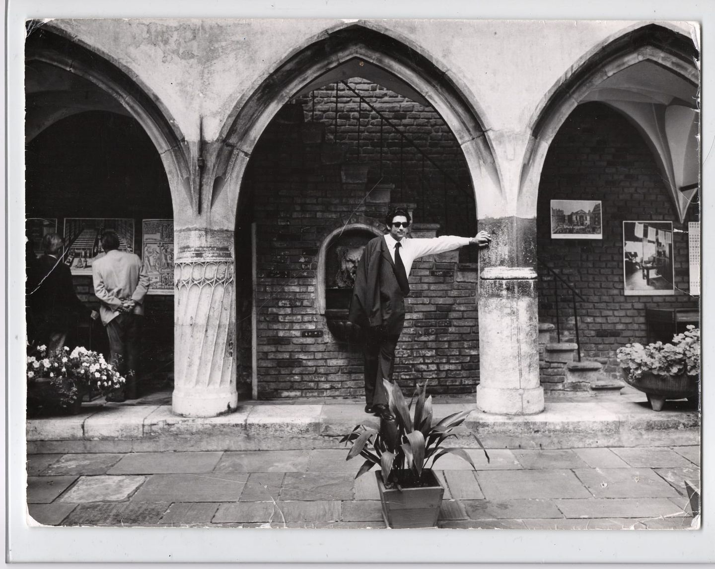 Black-and-white photo of a man in sunglasses leaning against a column in an arched stone courtyard.