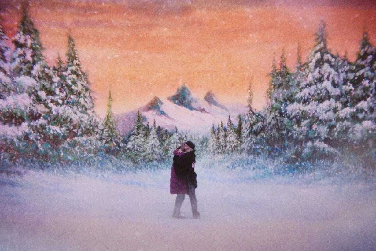 Two people embrace in a snowy forest landscape with mountains and a pink-orange sky in the background.