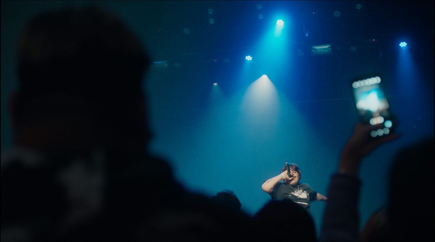 Performer singing into a microphone on a blue-lit stage, viewed from the audience.