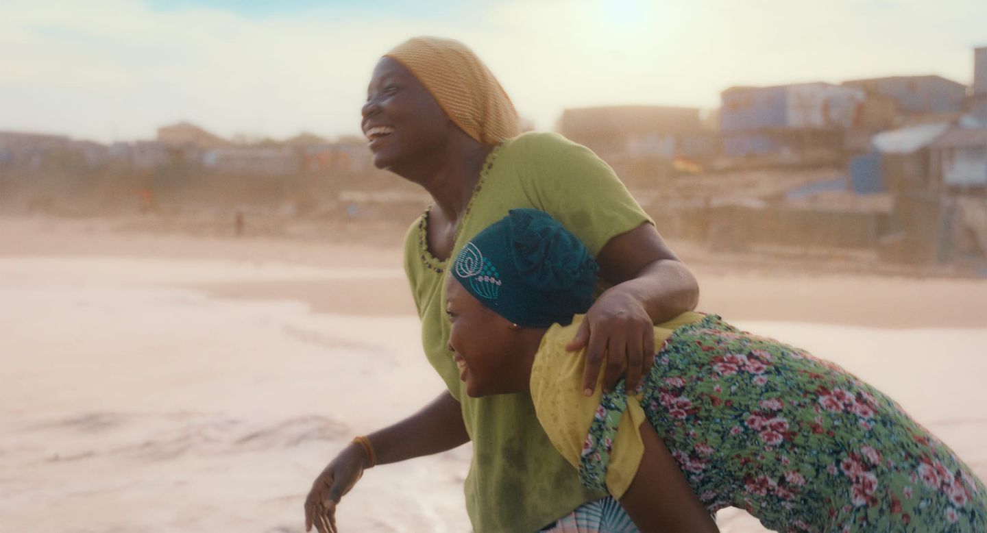 Two people in colourful clothing and headscarves laugh together on a sandy beach.