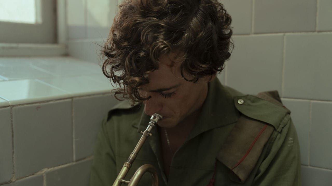 A curly-haired man in a green shirt plays a brass mouthpiece in a tiled room.