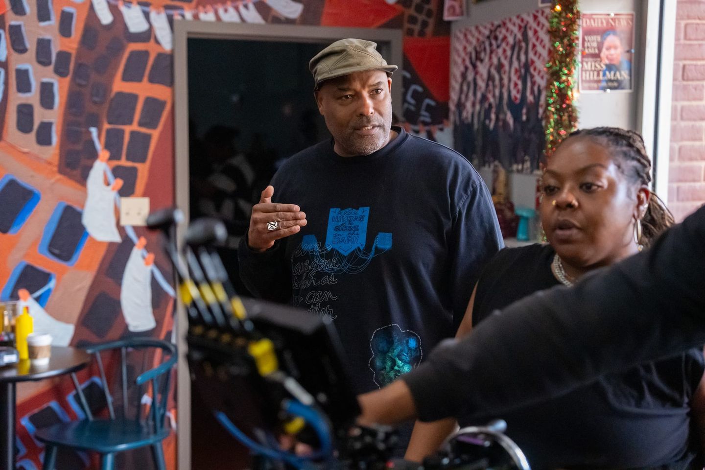 A man in a cap gestures beside a woman in a colourful room, with camera equipment in the foreground.