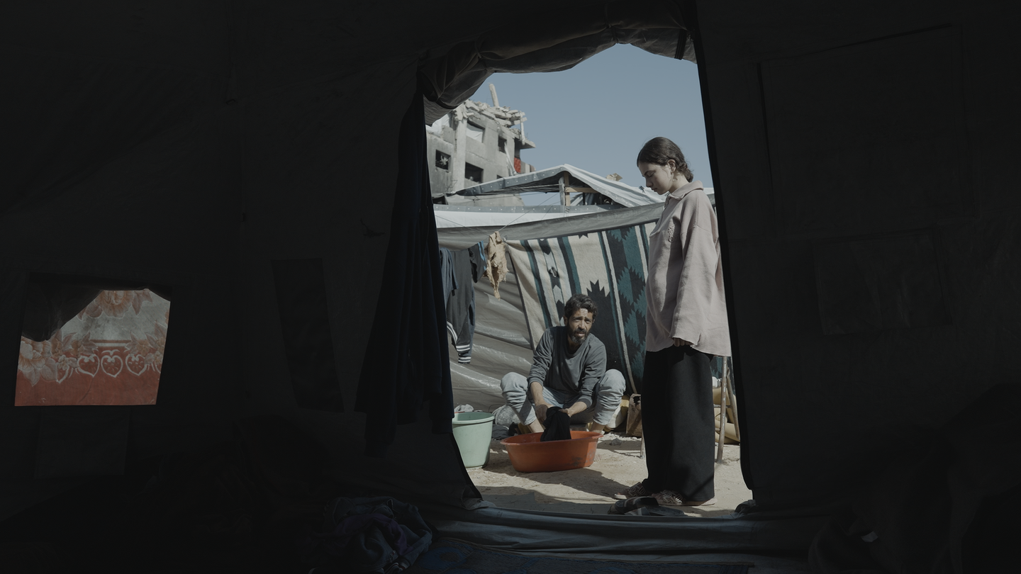 Inside a dark tent, a young person stands beside a crouching man washing clothes in a basin, with tarps and a damaged building behind them.