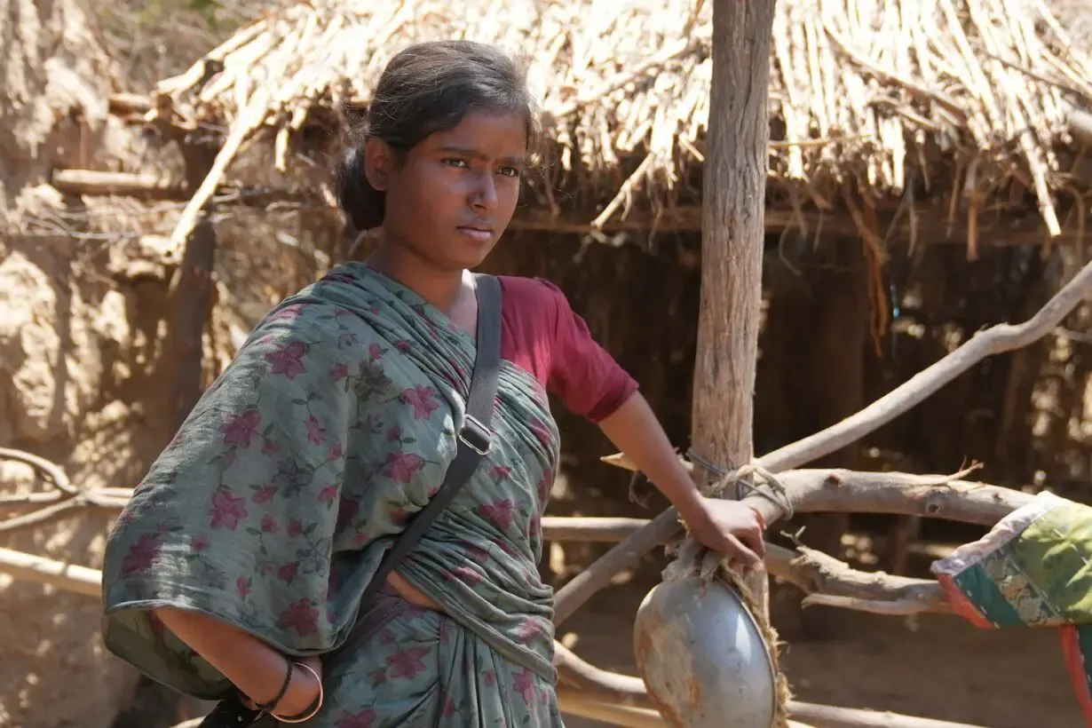 A young woman in a floral green sari stands beside a wooden fence in front of a thatched rural structure, holding a metal pot.