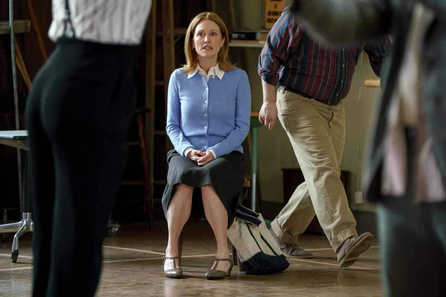 A woman in a blue cardigan sits with clasped hands in a room, surrounded by partially visible standing people.