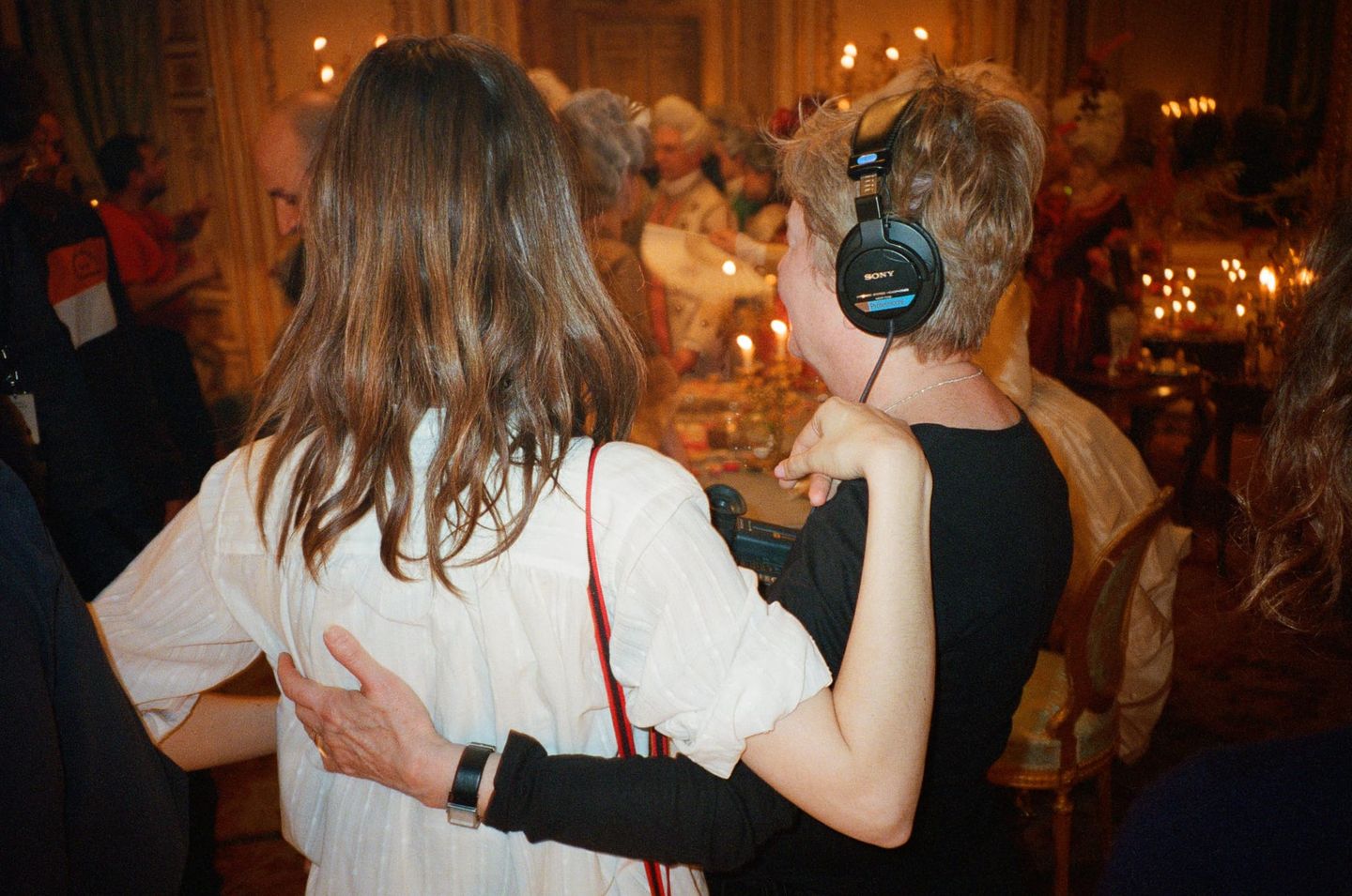 Two people seen from behind with arms around each other in a crowded, candlelit ornate room; one wears large headphones.