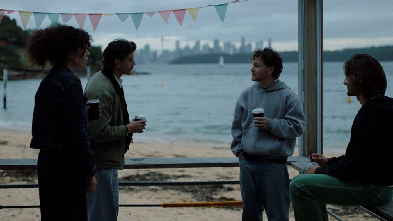 Four people holding takeaway coffee cups talk under bunting in a beachfront shelter, with water and a distant city skyline behind them.