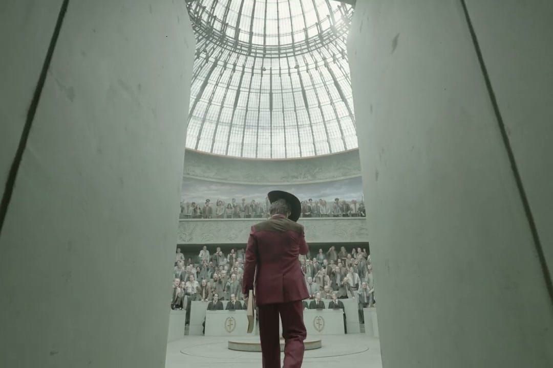 Person in a burgundy suit and black hat walks into a large domed auditorium filled with seated spectators.