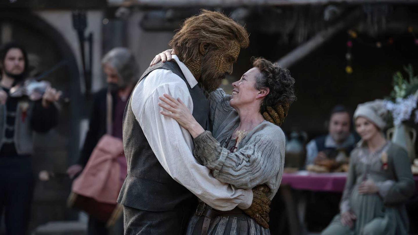A tall, hairy beast-like figure in a vest and white shirt embraces a smiling woman in period clothing, with musicians and seated guests blurred in the background.
