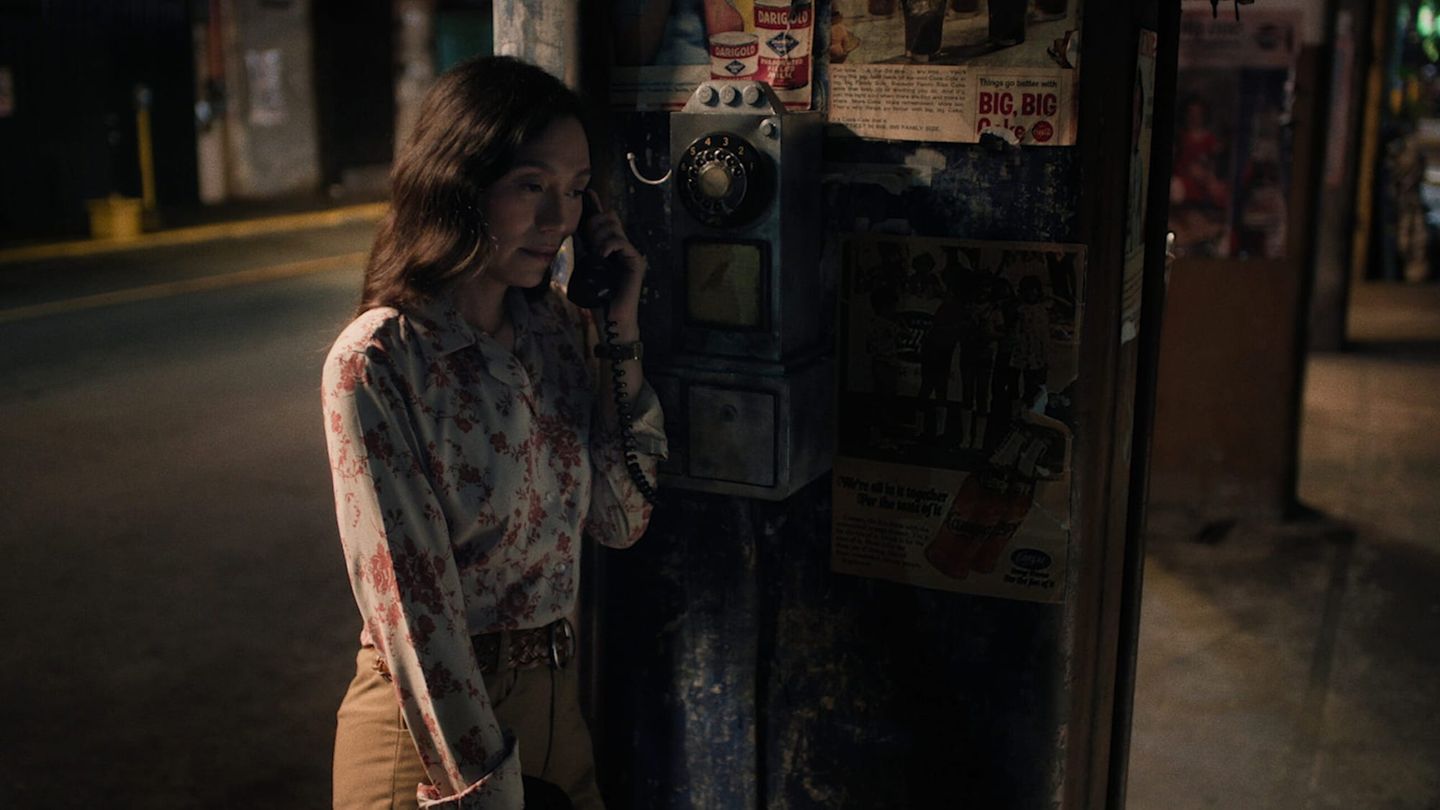 A woman in a white blouse with red floral prints and tan trousers uses a black rotary payphone in a dim, warm-toned street-side booth covered with faded advertisements.