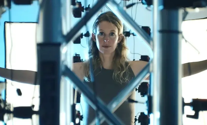 A woman stands behind a metal mechanical framework with crossed support bars and bright backlighting.