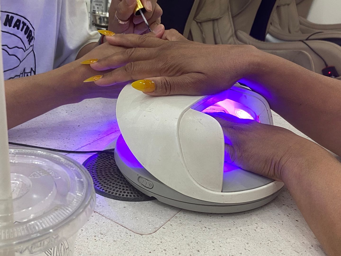 Nail technician with yellow nails applies polish to a client's hand while the other hand cures under a UV/LED nail lamp.
